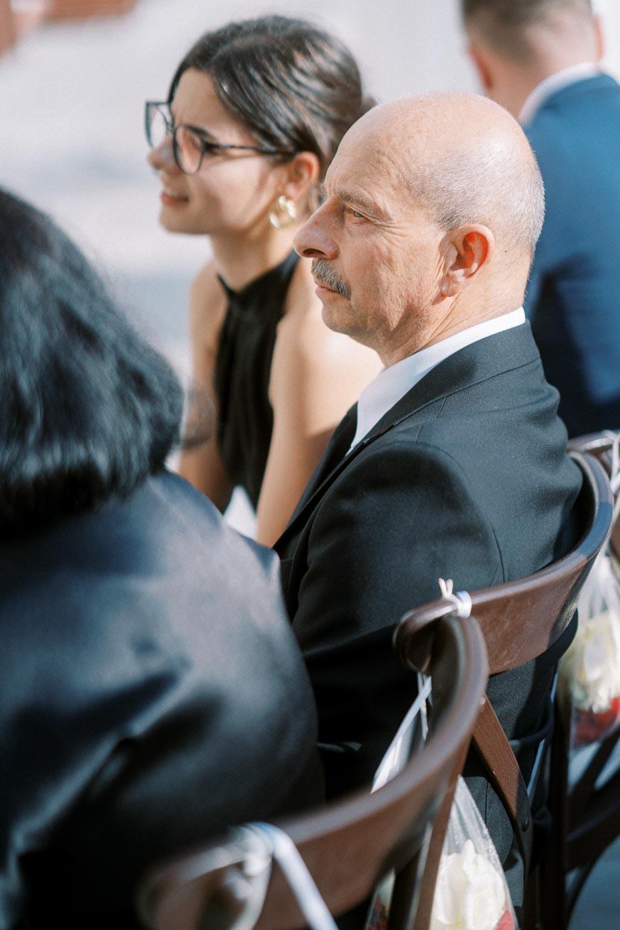 Guests attentively watching a ceremony, with a bald man in a suit and a woman wearing glasses seated on wooden chairs.
