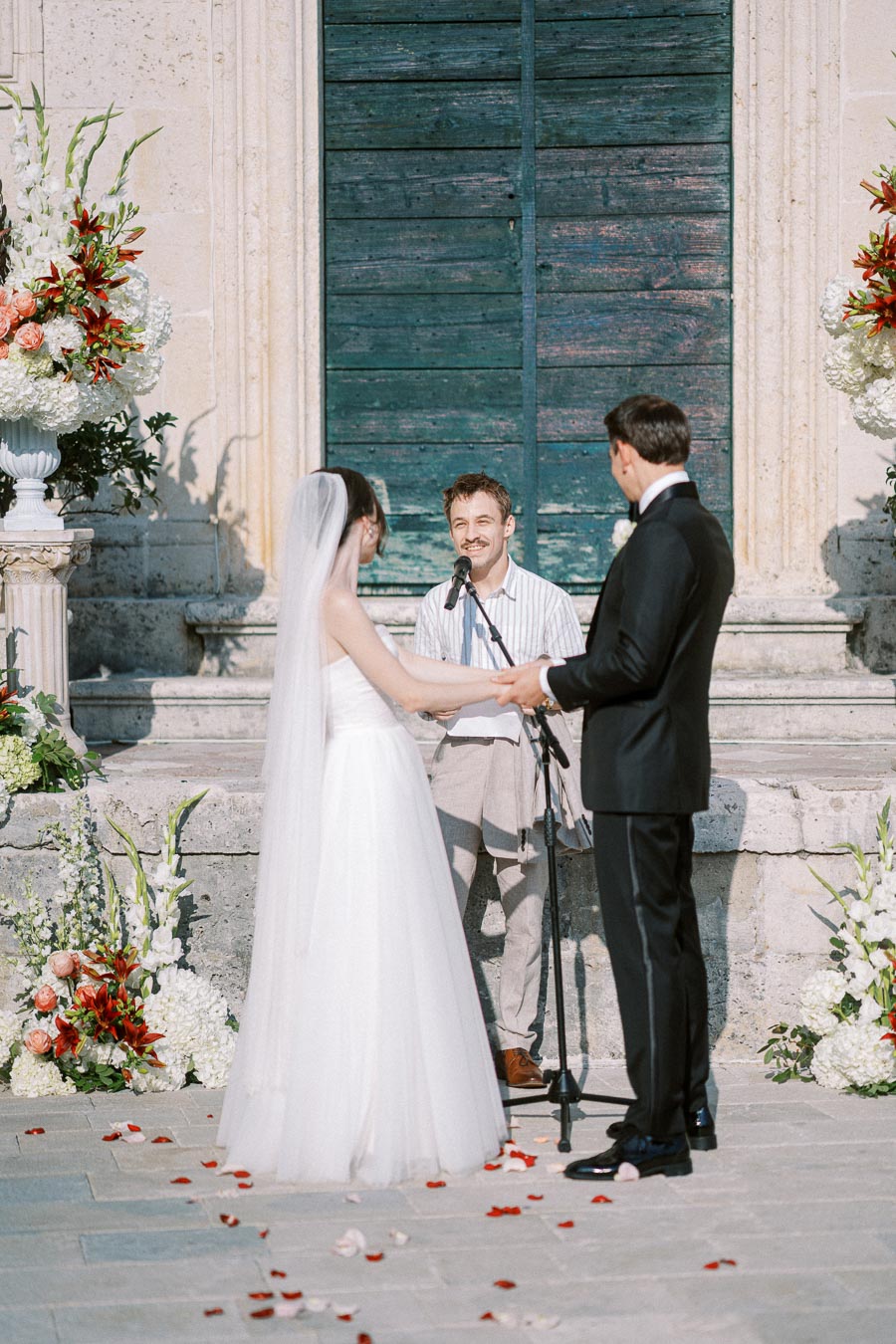 Elegant outdoor wedding ceremony with a couple holding hands in front of an officiant, surrounded by beautiful floral arrangements, and a rustic blue wooden backdrop.
