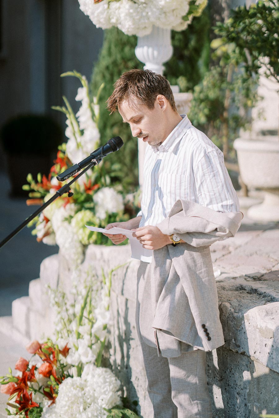 Man in a light-colored suit giving a speech at an outdoor event, standing at a microphone with floral arrangements in the background.