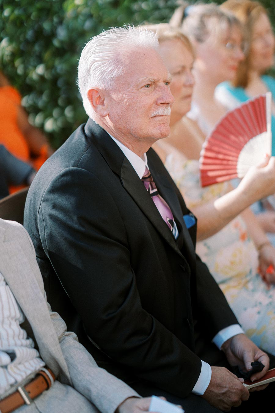 Elderly man in a formal suit sitting outdoors among a group of people.