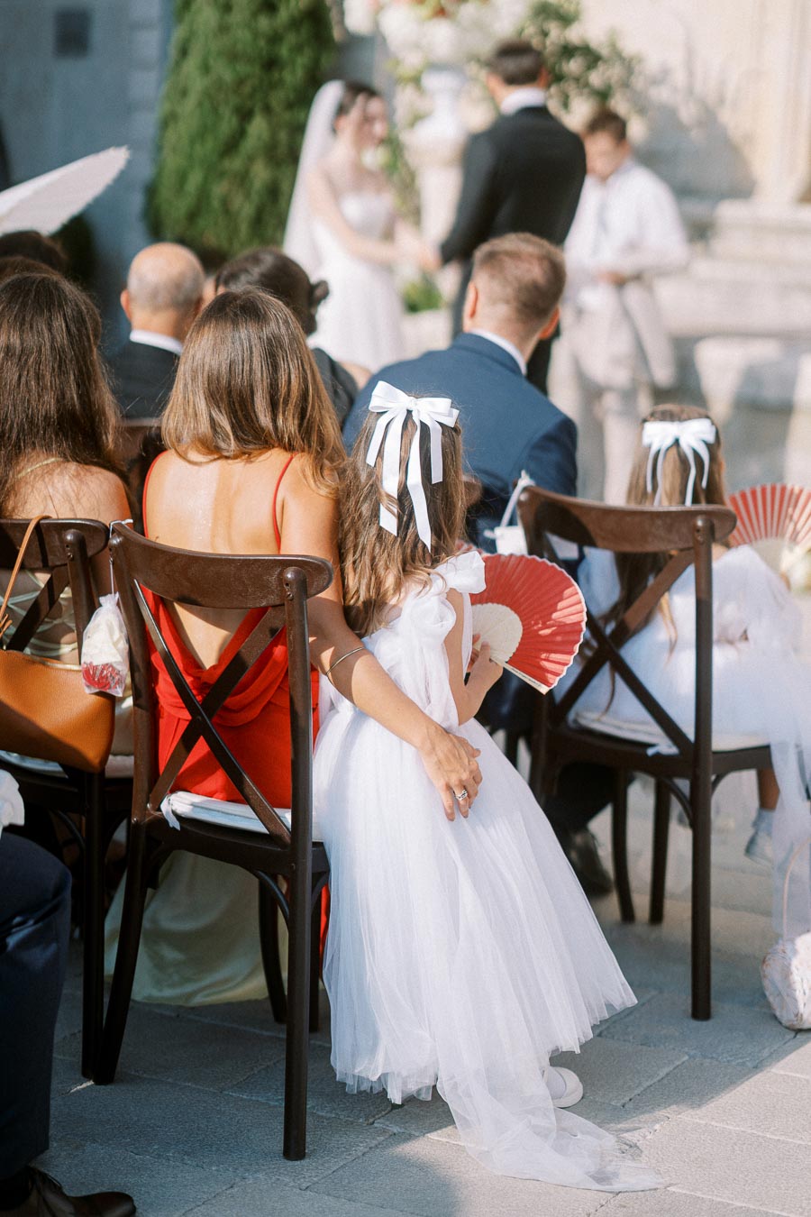 Outdoor wedding ceremony with guests seated, including a woman in a red dress and a child in a white dress holding a red fan. Bride and groom in the background exchanging vows.