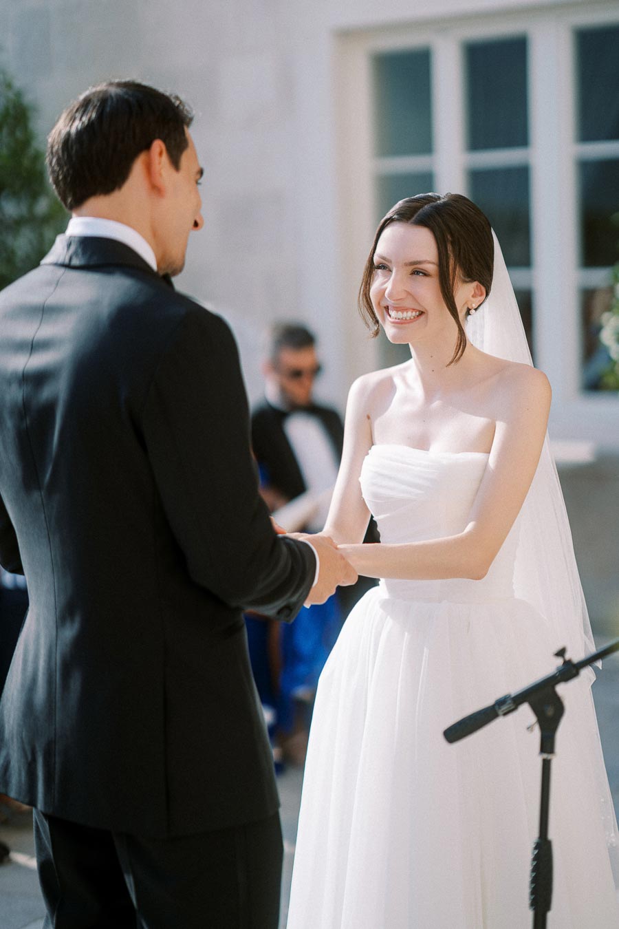 A bride and groom exchange vows at an outdoor wedding ceremony, both dressed in formal attire. The bride is wearing a white wedding dress and veil, smiling joyfully as she holds the groom’s hands.