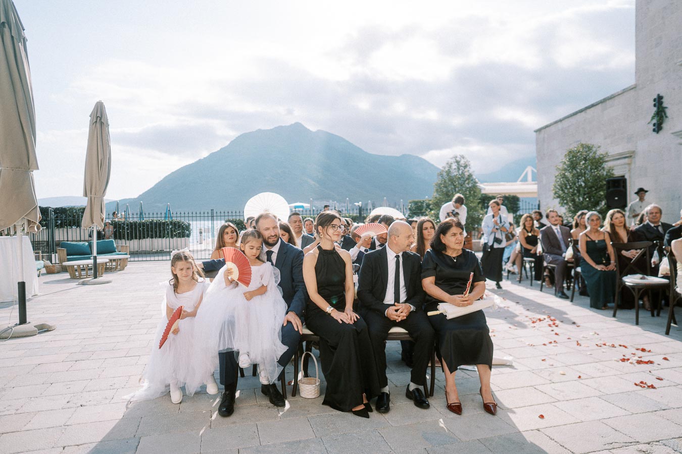 Outdoor wedding ceremony with guests seated in formal attire, mountains and sky in the background, featuring flower petals on the ground.