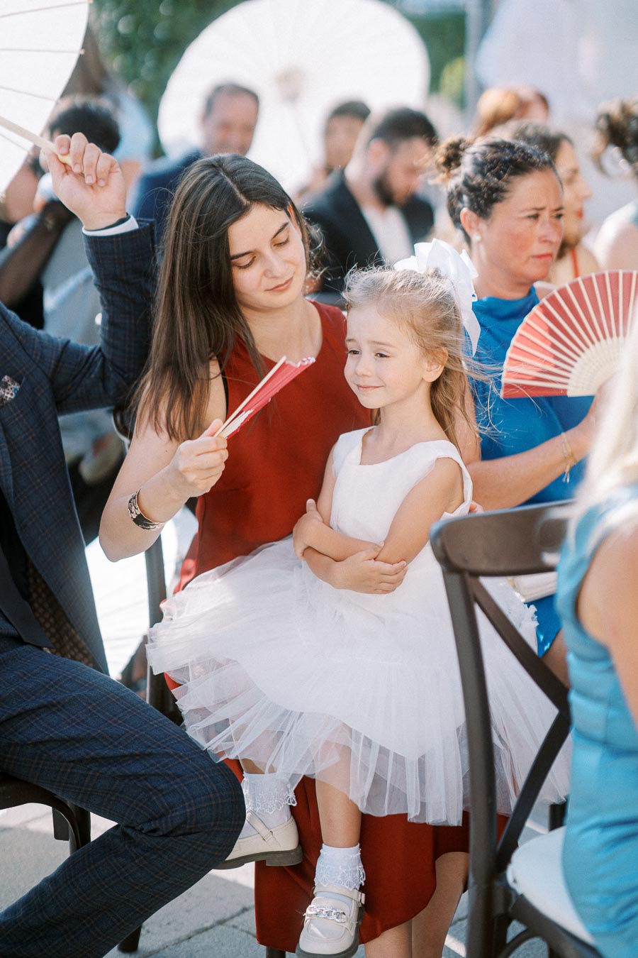 Woman in red dress holding a fan and smiling with a young girl in a white dress on her lap at an outdoor event, surrounded by people with parasols.