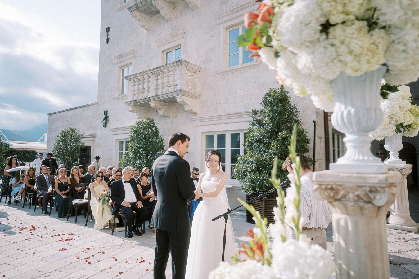 Outdoor wedding ceremony with a bride and groom standing in front of a beautifully decorated stone building, surrounded by guests seated on the patio. Elegant floral arrangements and clear skies enhance the romantic atmosphere.