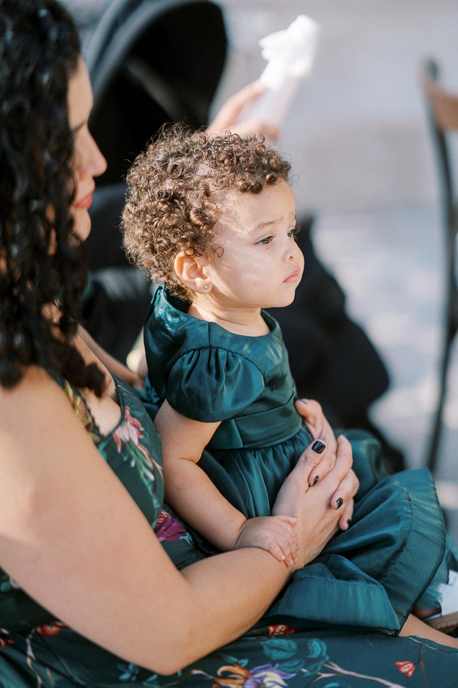 A mother holding her young child in her lap, both wearing matching green dresses, at an outdoor event.