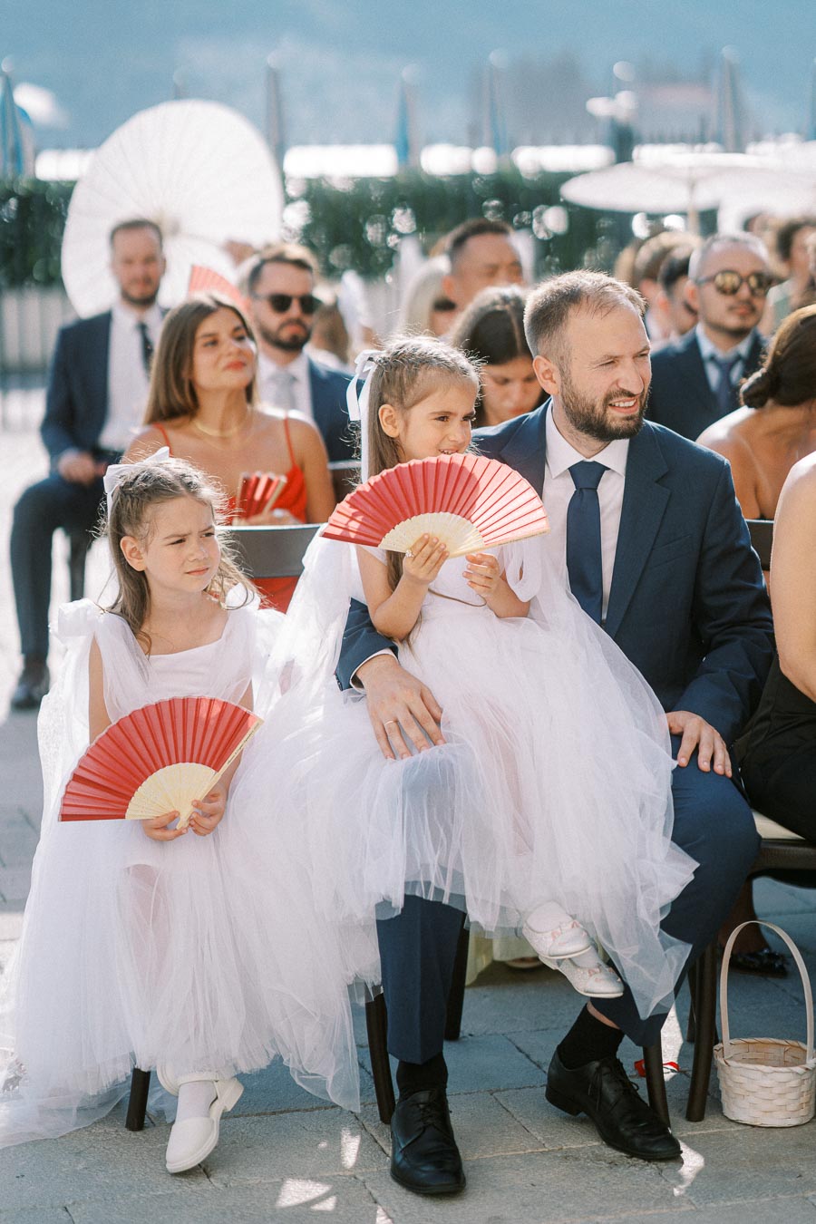 A well-dressed man in a blue suit sits with two young girls wearing white dresses and holding red fans at an outdoor wedding ceremony, surrounded by other elegantly dressed guests on a sunny day.