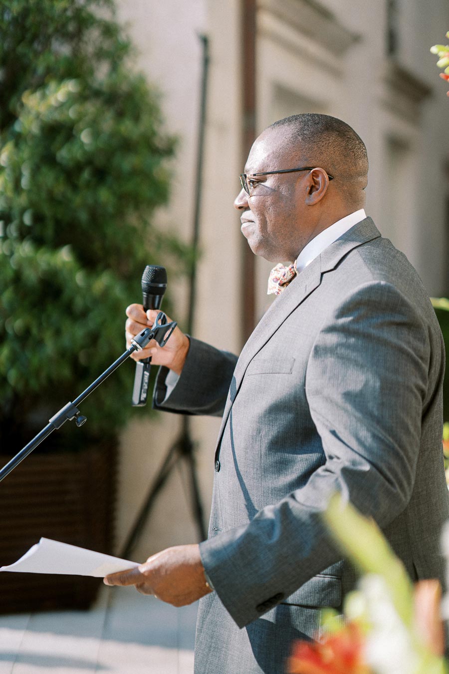 A man in a suit speaks into a microphone outdoors during a formal event, holding papers.