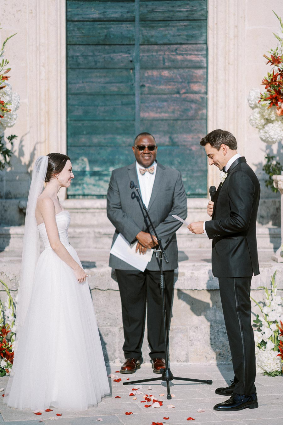 Bride and groom exchanging vows during an outdoor wedding ceremony, officiated by a man in a suit, with flower arrangements and a rustic backdrop.
