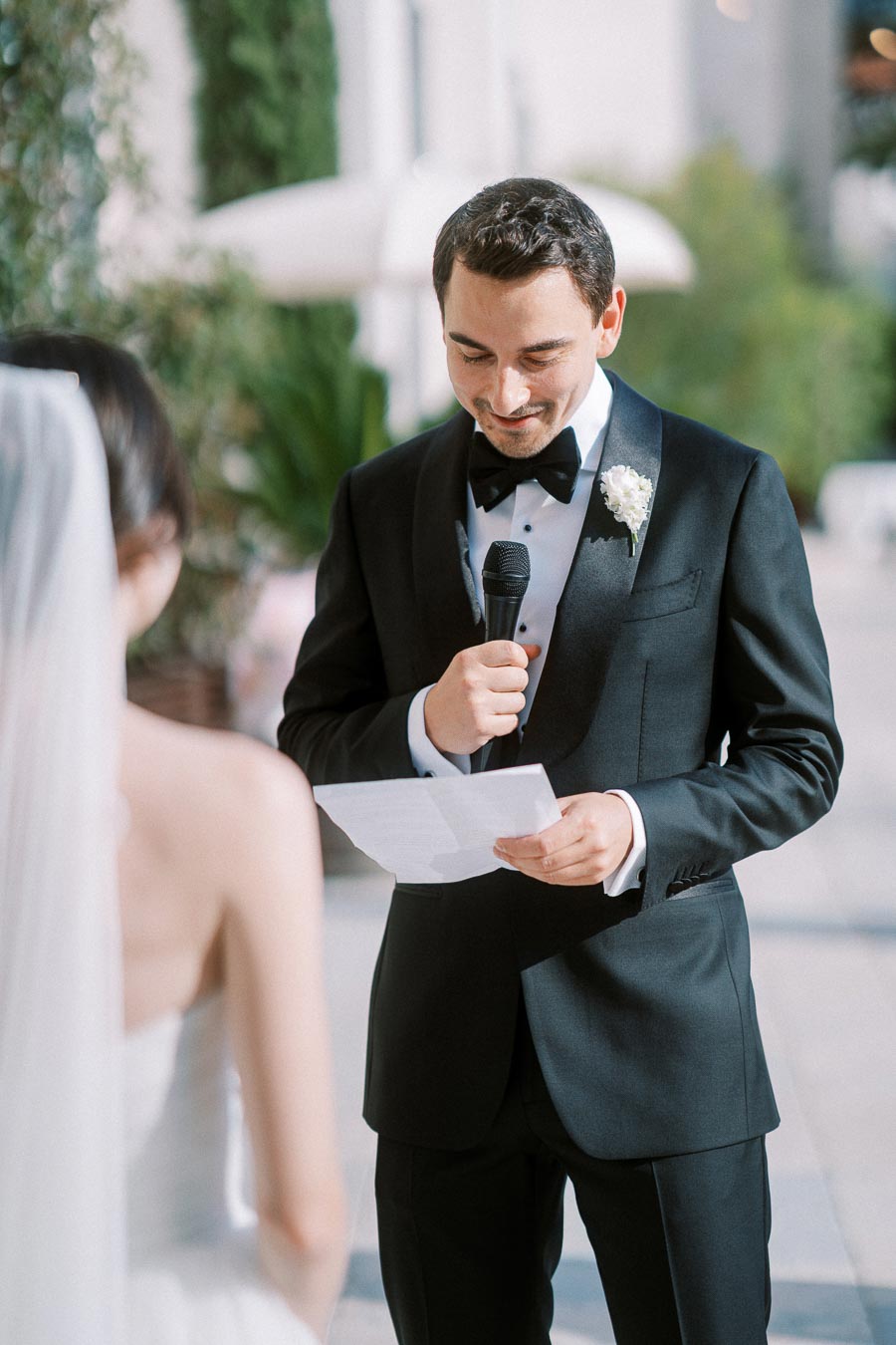A groom in a black tuxedo reads vows to the bride during an outdoor wedding ceremony.