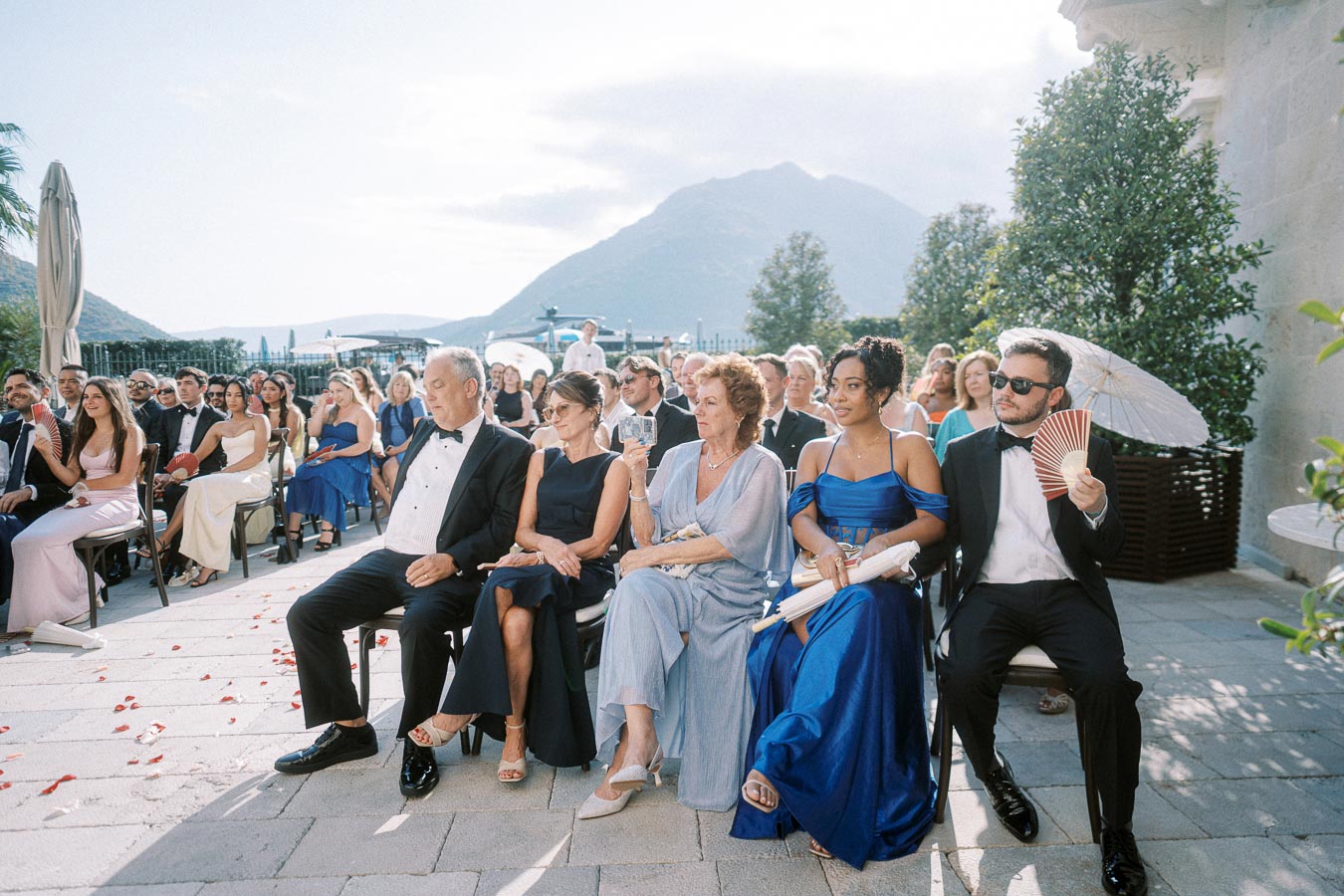 A group of elegantly dressed guests seated outdoors at a wedding ceremony with a scenic mountain view in the background.