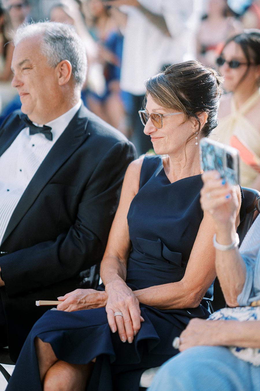 A couple dressed elegantly, seated at an outdoor event. The woman is wearing a blue dress and glasses, while the man is in a tuxedo. They appear engaged with the ceremony.