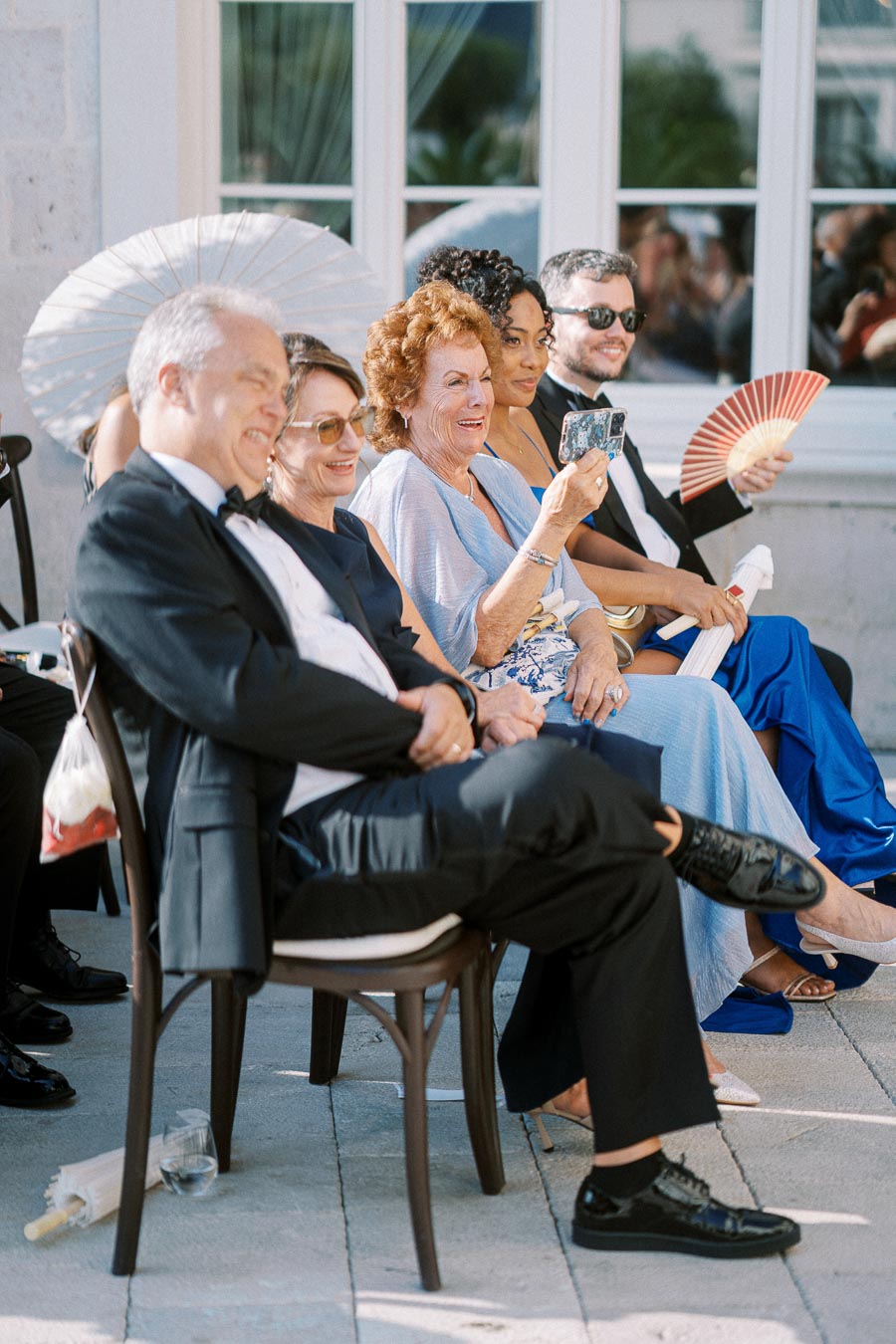 Group of elegantly dressed guests smiling and enjoying a wedding ceremony outside, with some holding paper fans for shade and comfort.