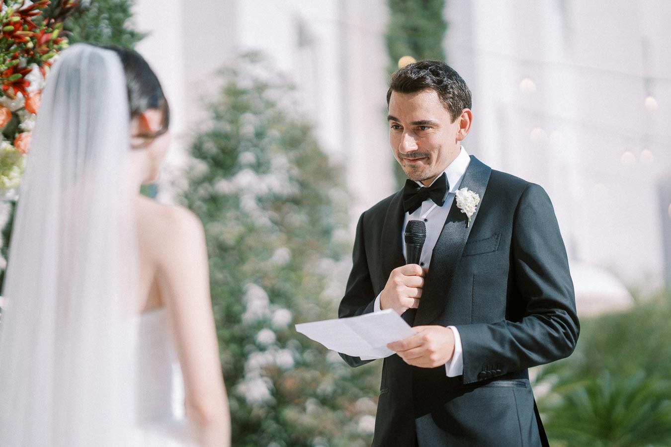 Groom in a black tuxedo holding a microphone reads vows to the bride in a white dress and veil, during an outdoor wedding ceremony with floral decorations.