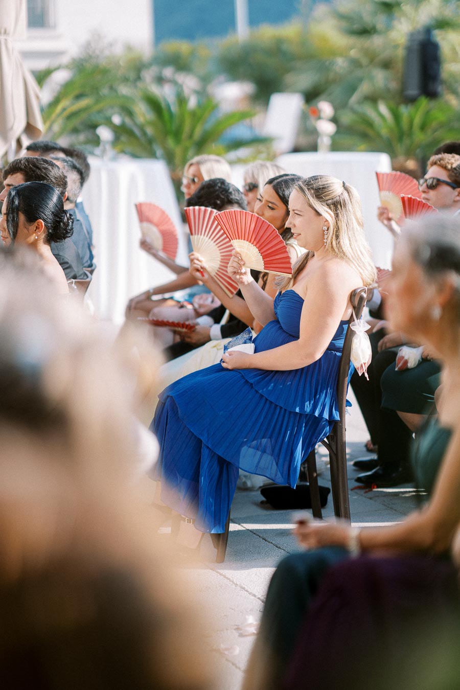 Guests seated outdoors at a wedding ceremony, dressed elegantly in colorful attire, with some holding red fans, surrounded by lush greenery.