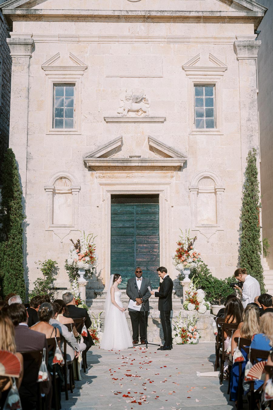 Outdoor wedding ceremony in front of a historic stone church with large wooden doors, featuring a couple exchanging vows, officiant, and guests seated on either side. Elegant floral arrangements and scattered rose petals enhance the romantic setting.