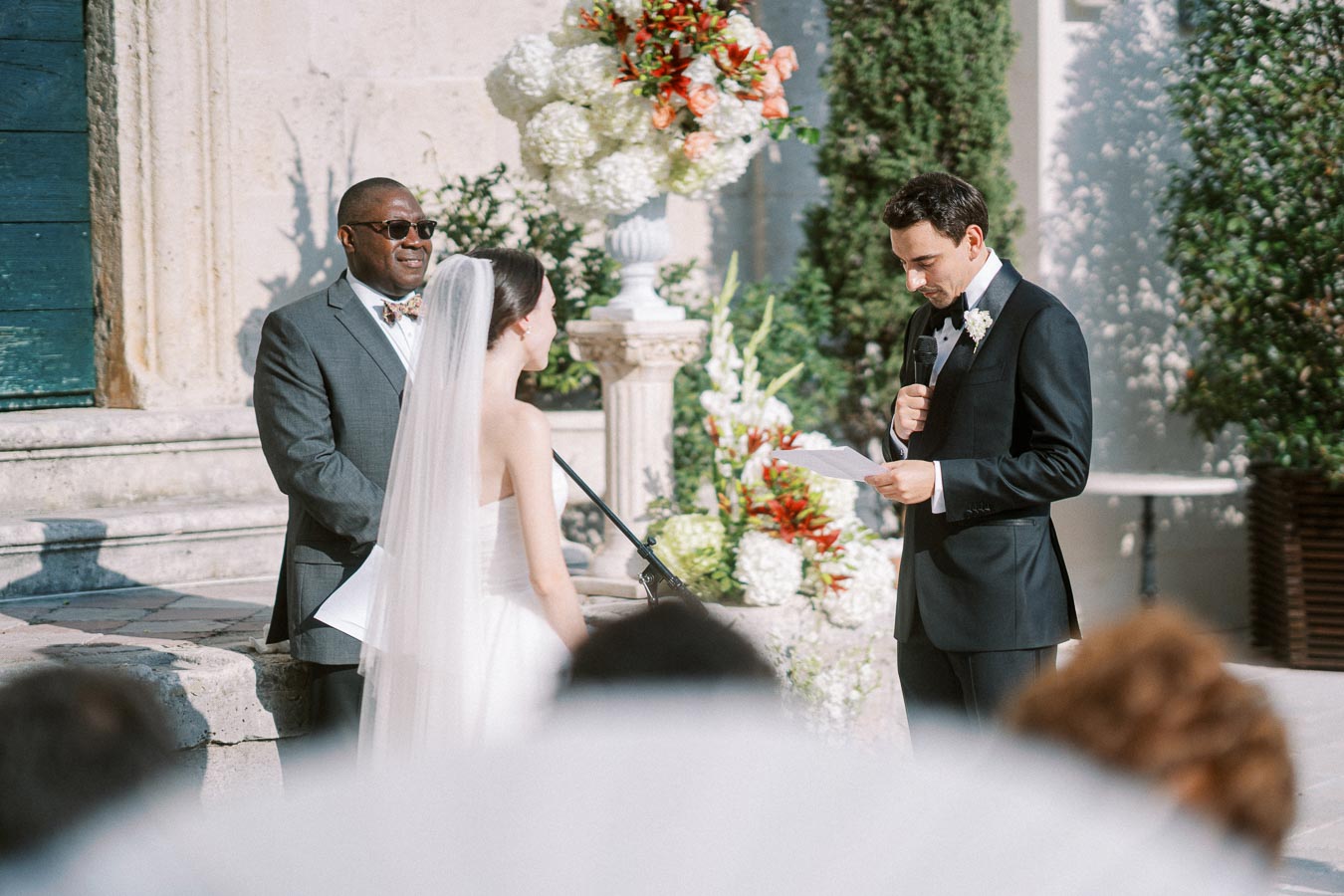 Outdoor wedding ceremony with a bride in a white gown and veil, a groom in a black tuxedo reading vows, and an officiant in sunglasses and a gray suit. Floral arrangements and greenery decorate the setting.