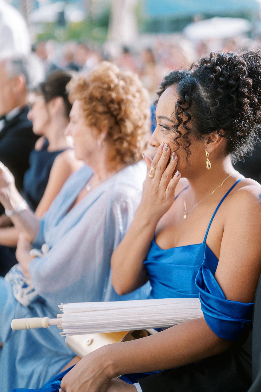 Audience in elegant attire at an outdoor event, featuring a woman in a vibrant blue dress holding a parasol, expressing surprise or delight.