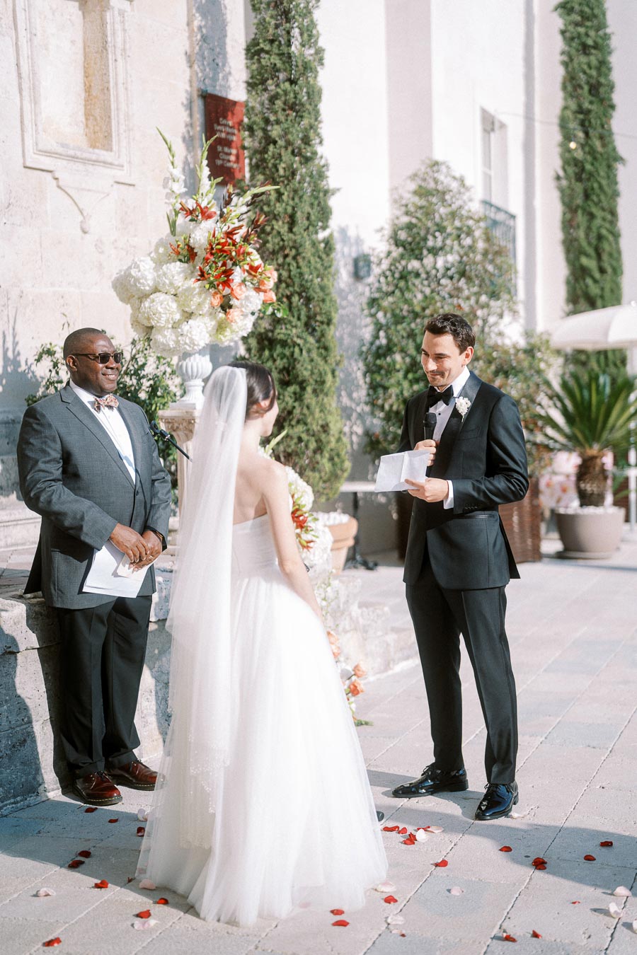 Outdoor wedding ceremony with groom in black suit reading vows to bride in white gown, with officiant and floral decorations.