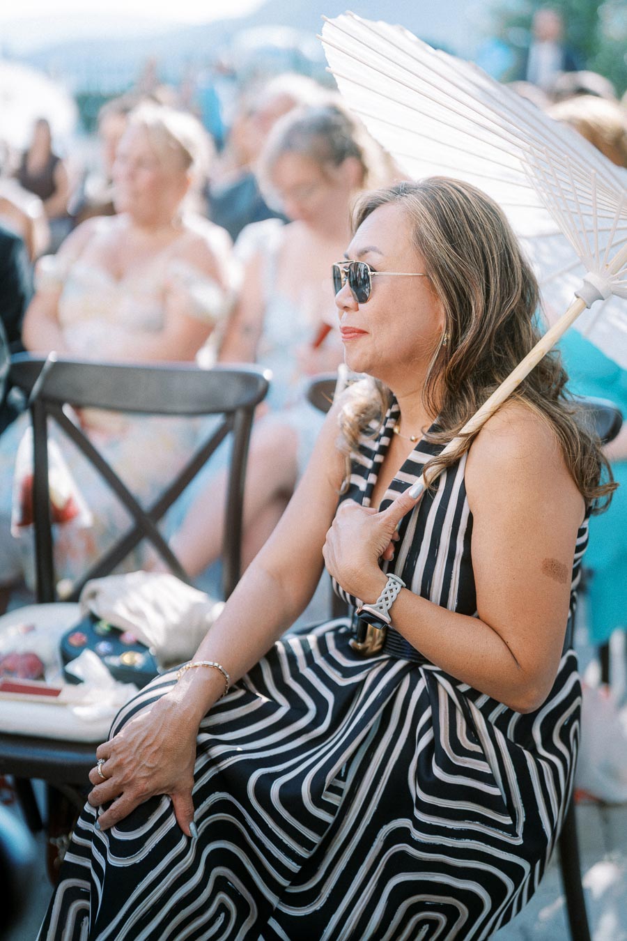 Woman in stylish striped dress, holding a white parasol and wearing sunglasses, sitting outdoors at a social event on a sunny day.