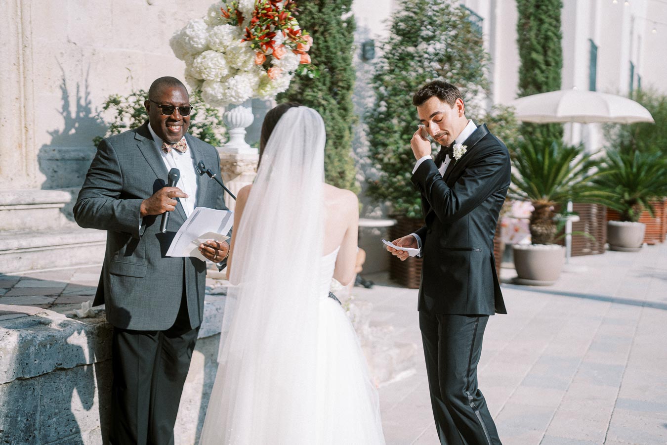 A bride and groom stand before a smiling officiant during an outdoor wedding ceremony, surrounded by lush greenery and elegant floral arrangements. The groom is seen wiping a tear while holding his vows, indicating the emotional significance of the moment.