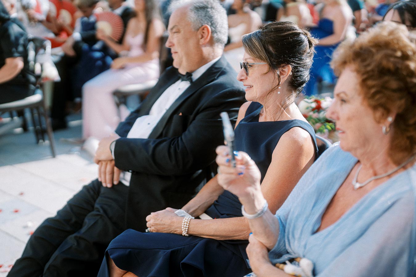 Three elegantly dressed guests seated at an outdoor event, with the woman at the center holding a small fan. The scene captures a formal gathering atmosphere, likely a wedding or a ceremony, with several attendees visible in the background.