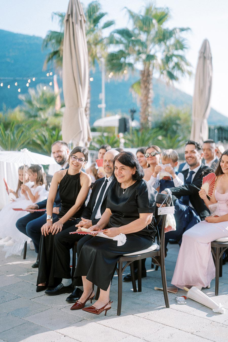 A group of well-dressed guests sitting in an outdoor setting at a wedding ceremony, surrounded by tropical plants and mountains in the background, enjoying the sunny weather with handheld fans.
