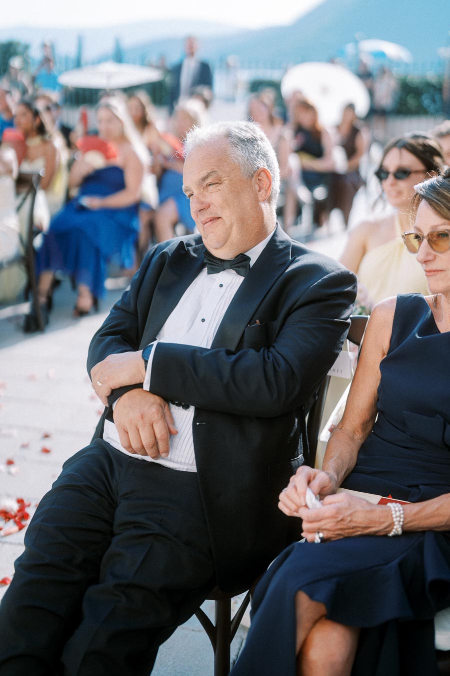 Elegant man in tuxedo seated outdoors, attending a formal event, surrounded by a crowd in elegant attire with mountain views in the background.