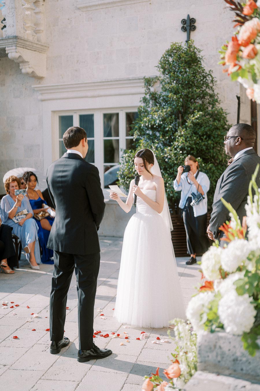 Outdoor wedding ceremony with bride in white dress reading vows to groom in black suit, surrounded by guests and floral decorations.