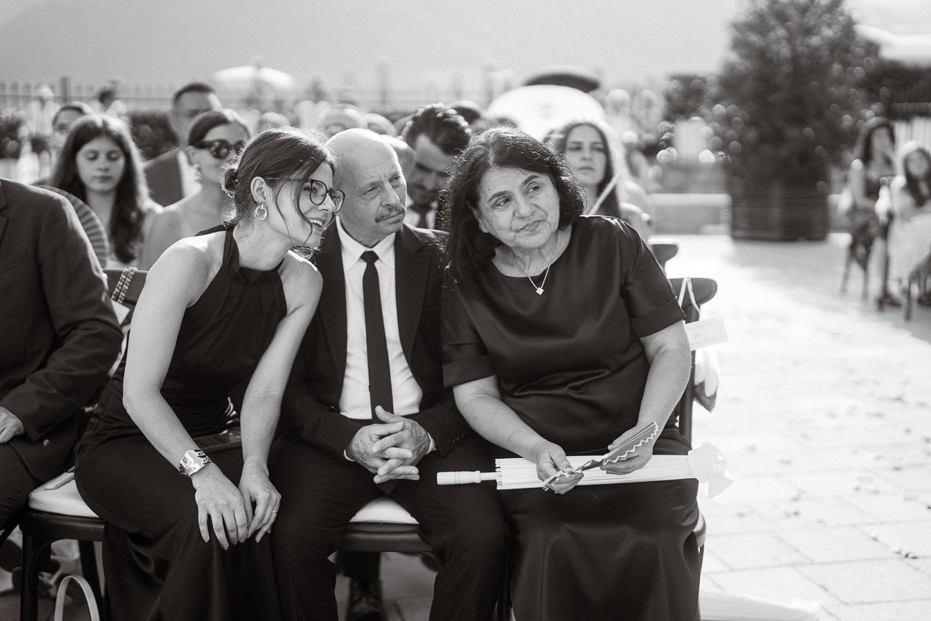 Black and white image of wedding guests sitting outdoors, including two women and a man in formal attire, with one woman leaning in to speak quietly.