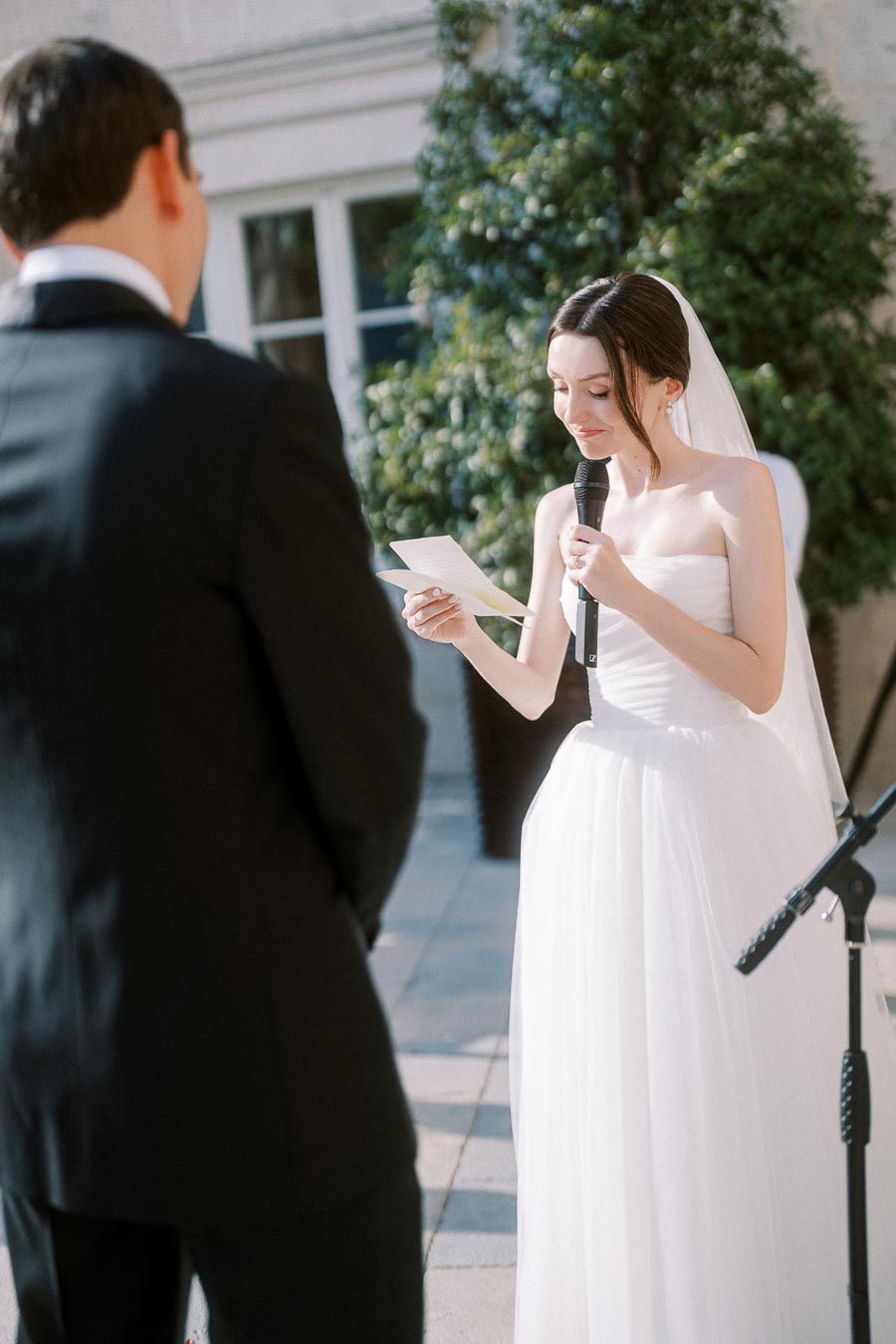Bride in white wedding dress delivering emotional vows outdoors, holding a microphone and paper, while groom in black suit listens attentively, with greenery in the background.