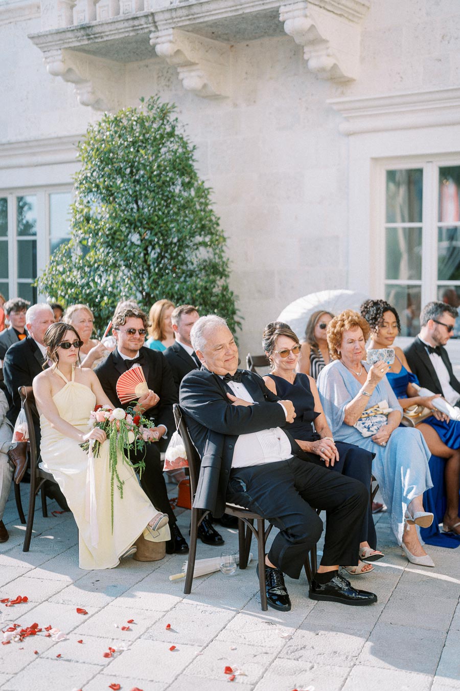Group of wedding guests seated outdoors, dressed in formal attire, at a sunlit ceremony.
