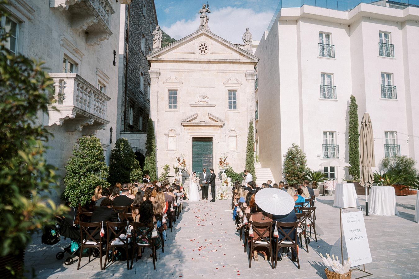 Outdoor wedding ceremony at a historical stone chapel with guests seated along a decorated aisle under a clear blue sky.