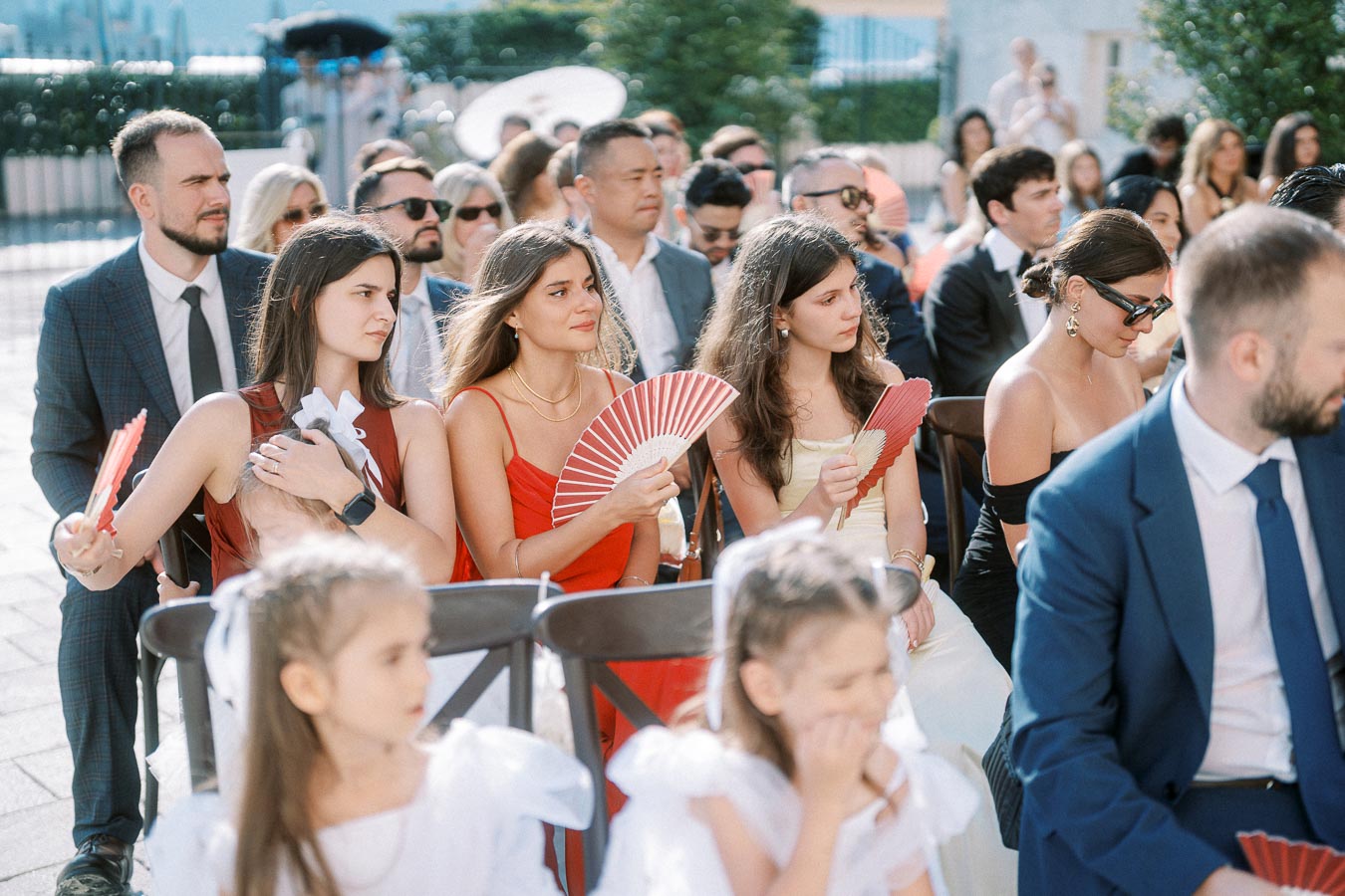 A diverse group of well-dressed guests seated outdoors at a formal event, holding red fans to stay cool under the sun.