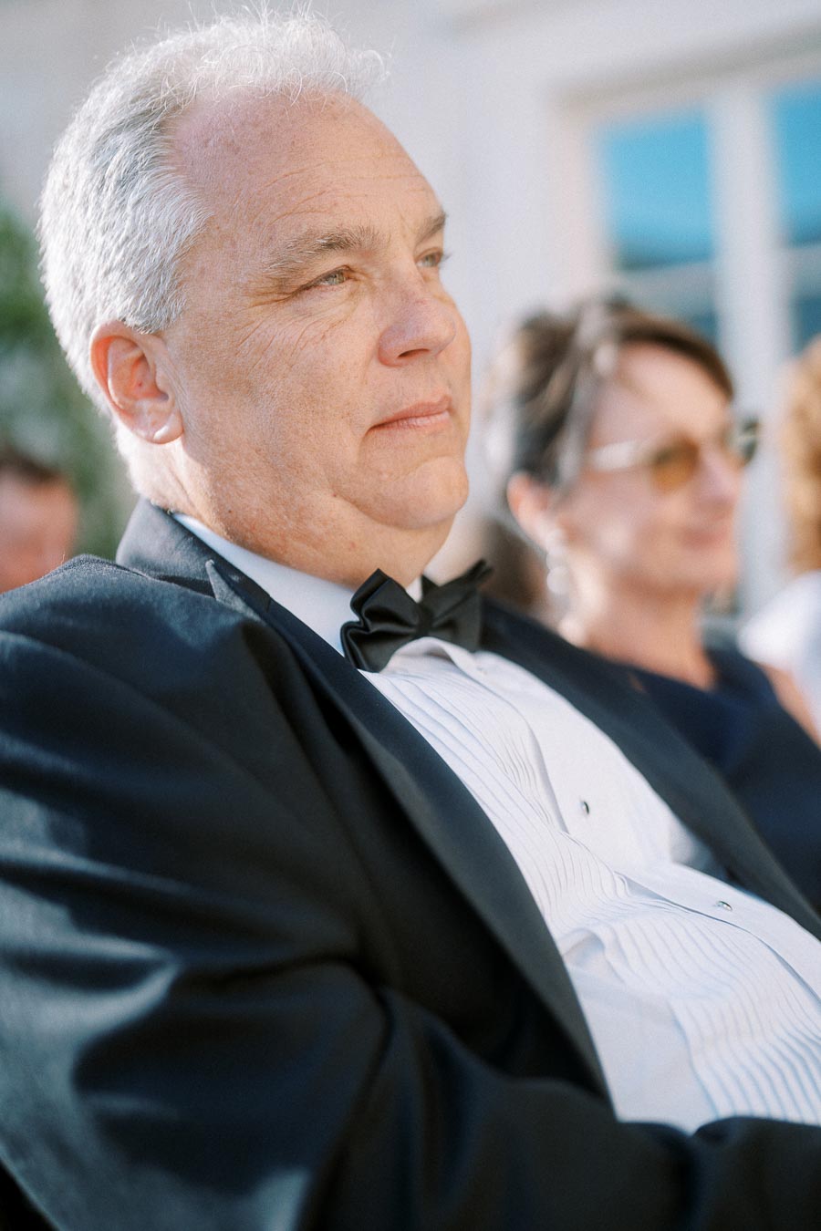 Elderly man in formal attire with a bow tie and suit, looking thoughtfully at an outdoor event.