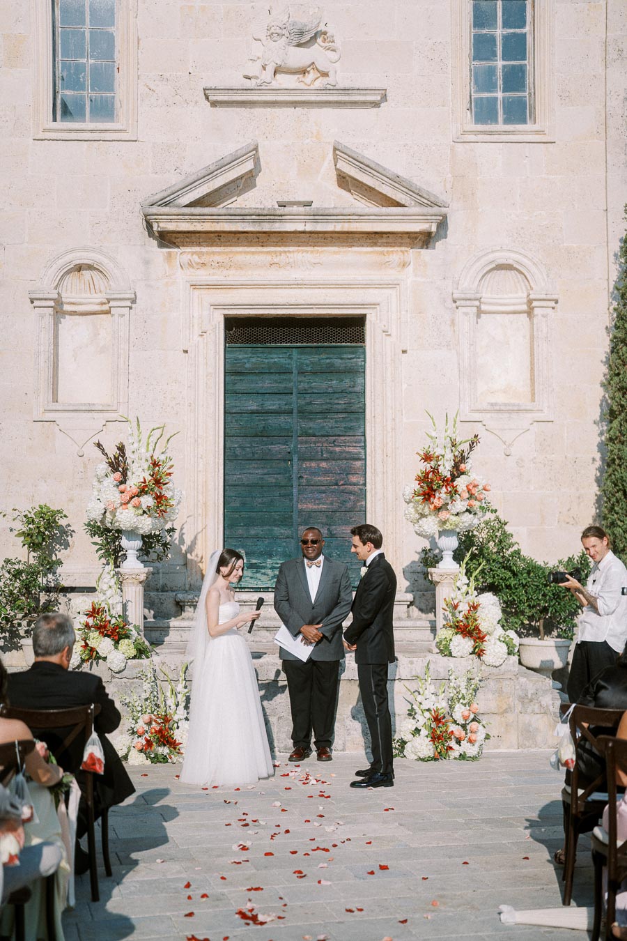 Outdoor wedding ceremony taking place in front of a historic stone building with elegant floral decorations, featuring a bride in a white gown holding a microphone, a groom in a black suit, and an officiant standing between them.