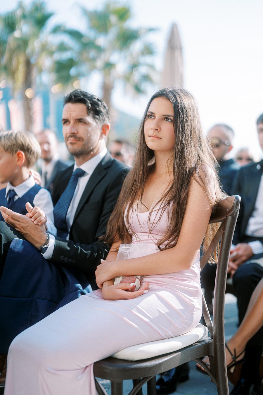 A young woman in an elegant pink dress sits attentively during an outdoor event, with a man in a suit and other attendees in the background, surrounded by a sunny setting with palm trees.