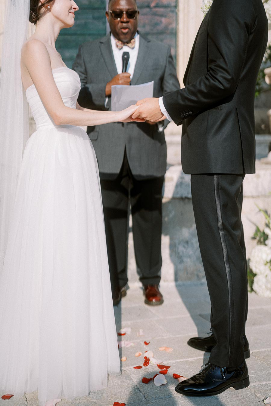 Wedding ceremony with bride and groom holding hands, officiant speaking, and rose petals scattered on the ground.