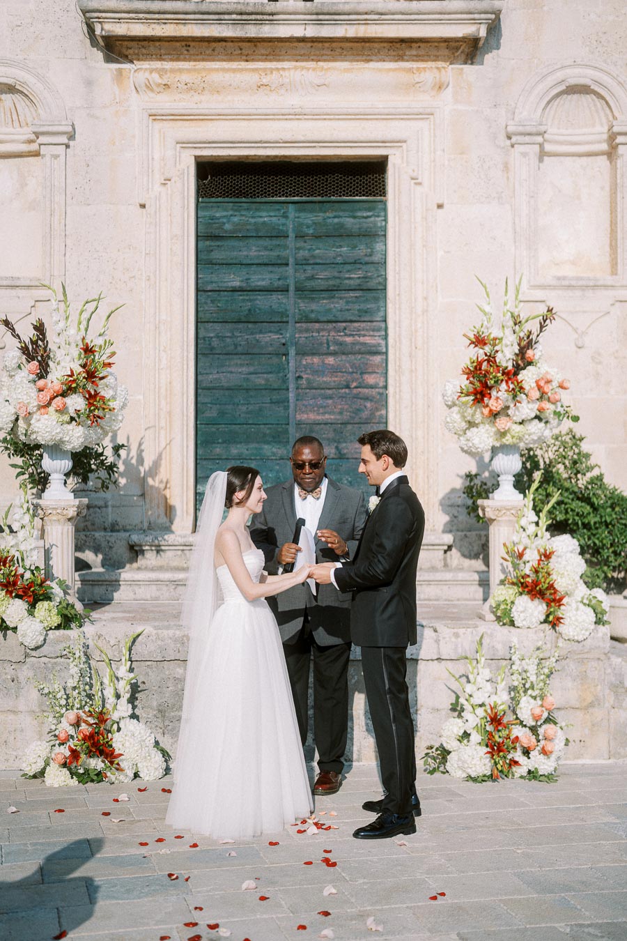 A bride and groom holding hands during an outdoor wedding ceremony in front of a historic stone building, surrounded by elegant floral arrangements.