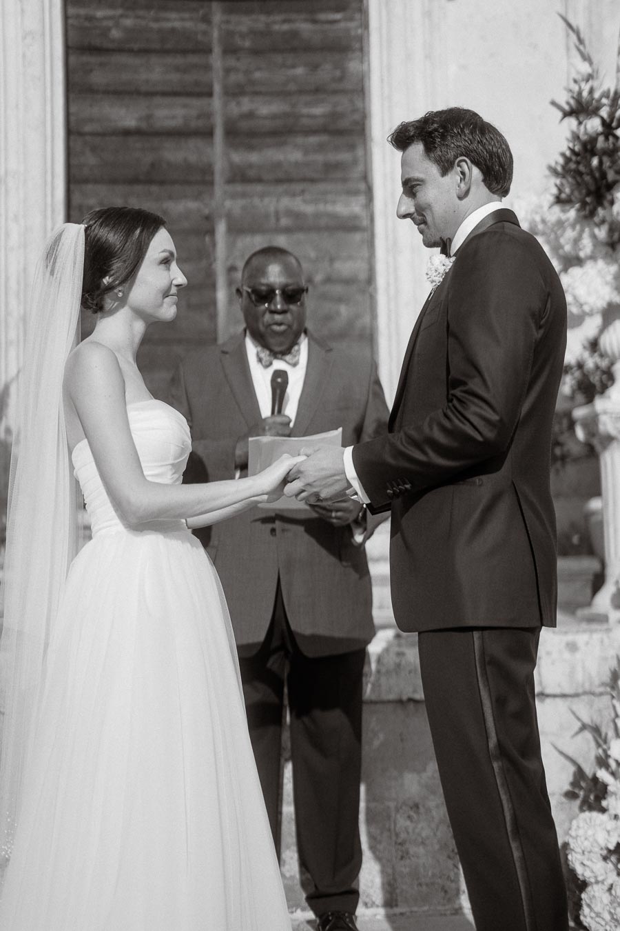 Black and white photograph of a wedding ceremony, featuring a bride and groom holding hands and exchanging vows in front of an officiant, with a historic wooden door backdrop.