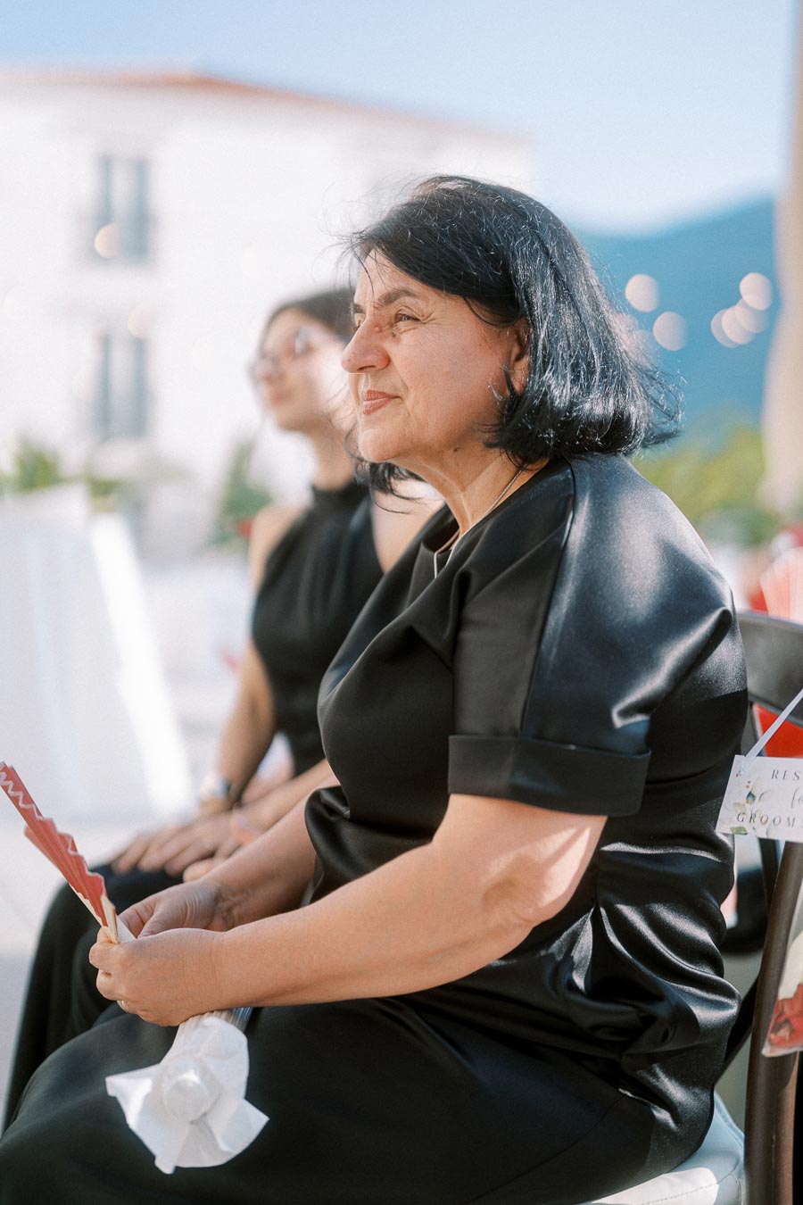 Elderly woman in black dress seated outdoors at a formal event, holding a red paper fan with a serene expression.