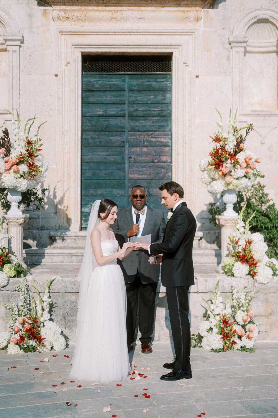 Bride and groom exchanging rings at an outdoor wedding ceremony with a stone backdrop and floral arrangements.