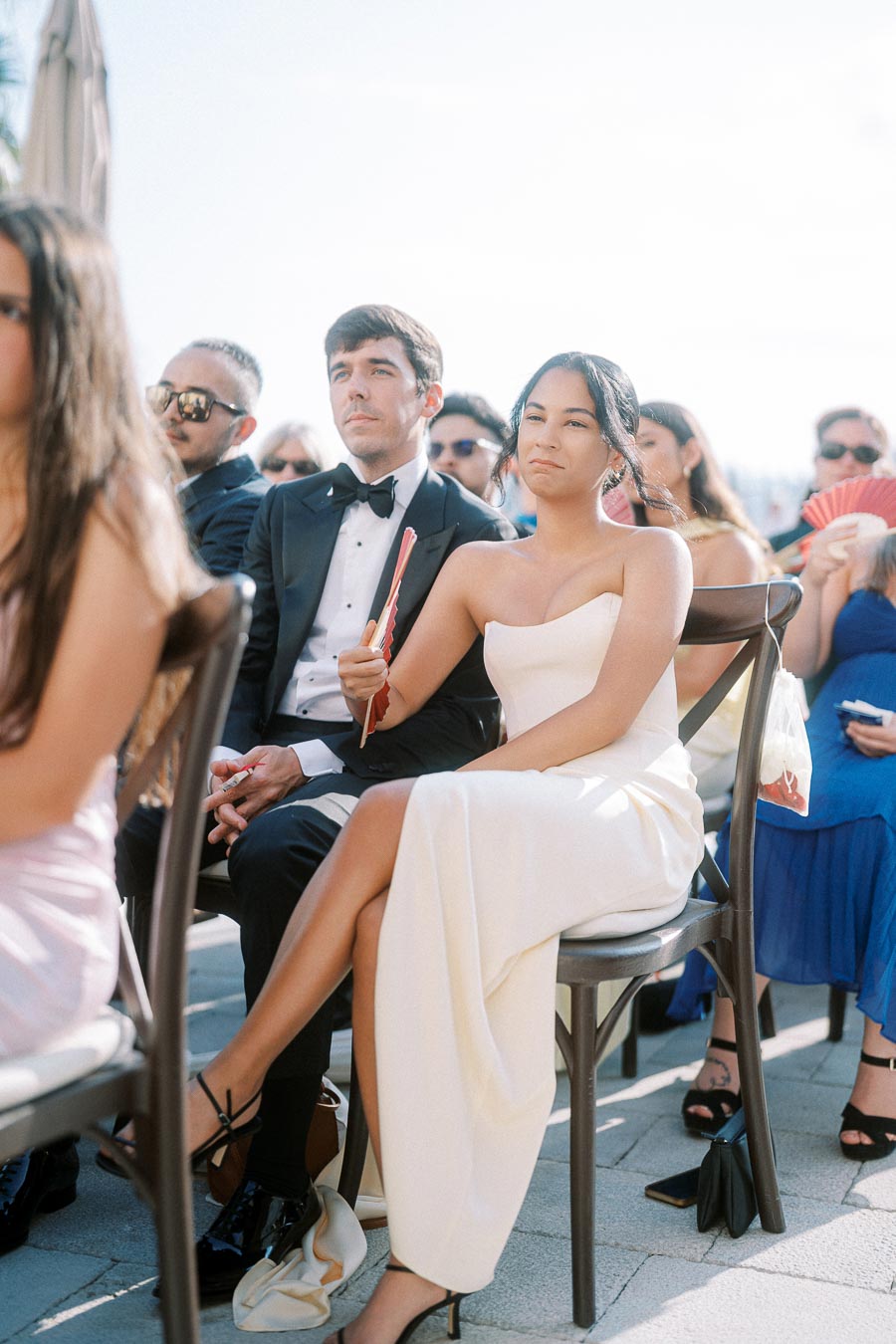 A formal outdoor event with guests seated, including a man in a tuxedo and a woman in a white dress holding a red fan.