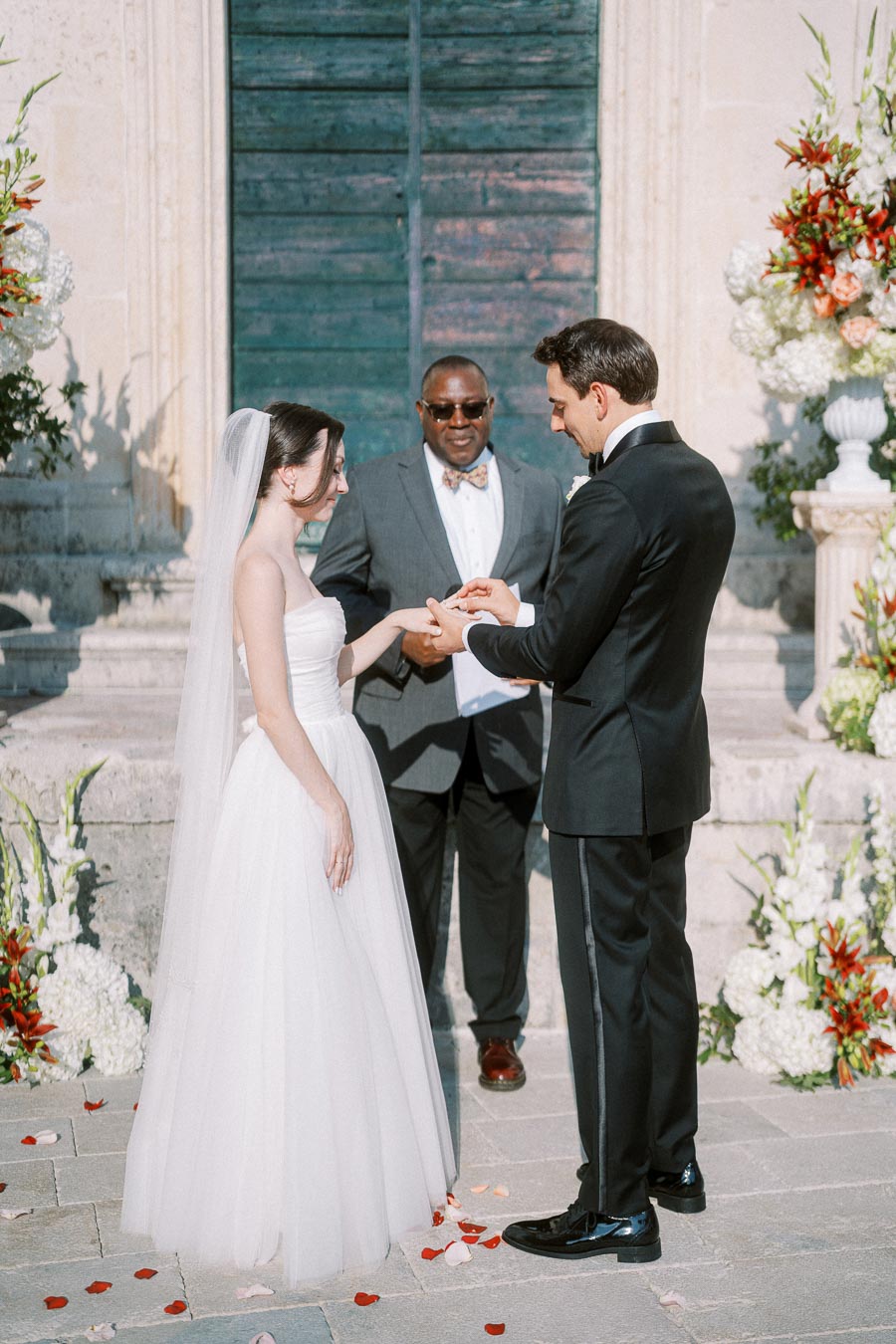 A bride and groom exchanging rings during an outdoor wedding ceremony, with an officiant presiding, surrounded by floral decorations and a historic stone backdrop.