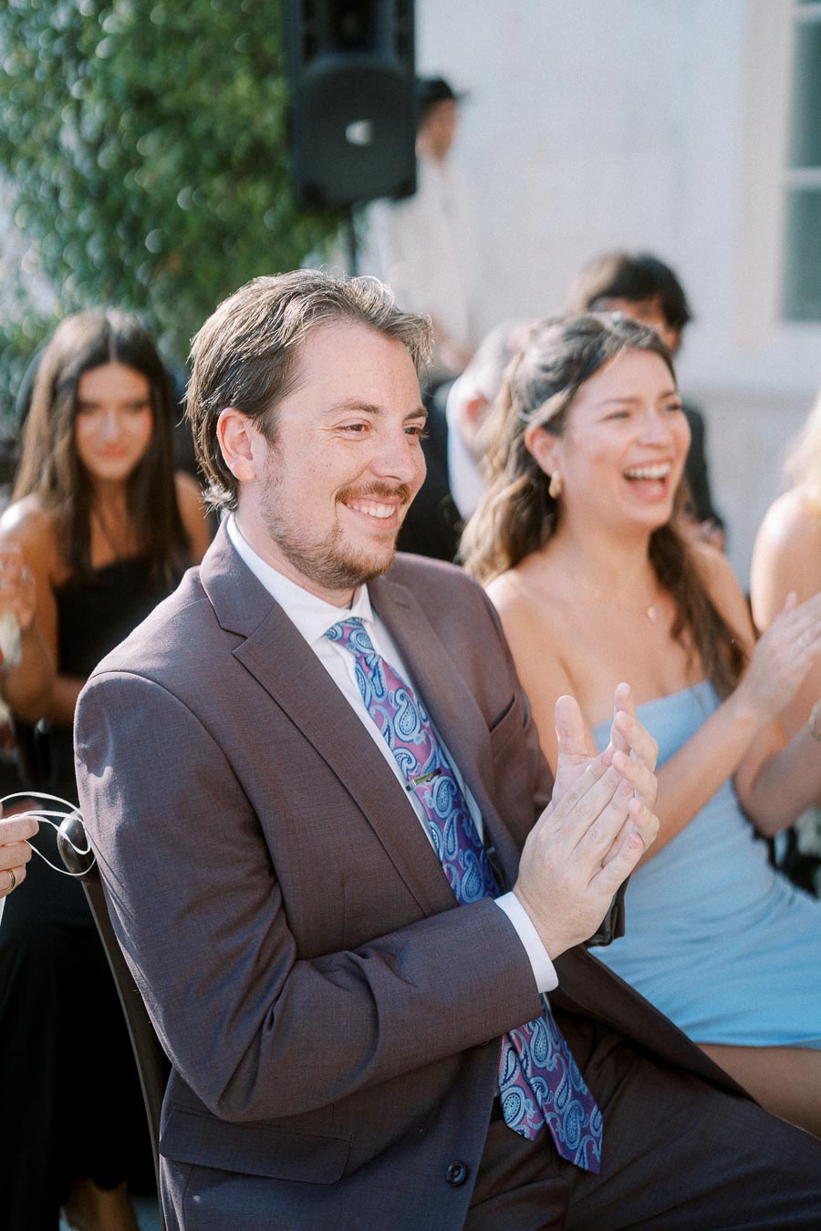 Man in a suit clapping and smiling at an outdoor event, with a woman in a blue dress clapping beside him.