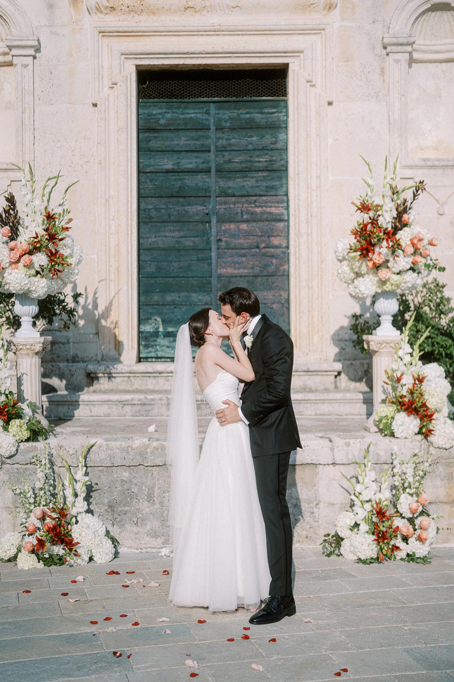 A bride and groom share a romantic kiss in front of a historic building, adorned with elegant floral arrangements, during an outdoor wedding ceremony.