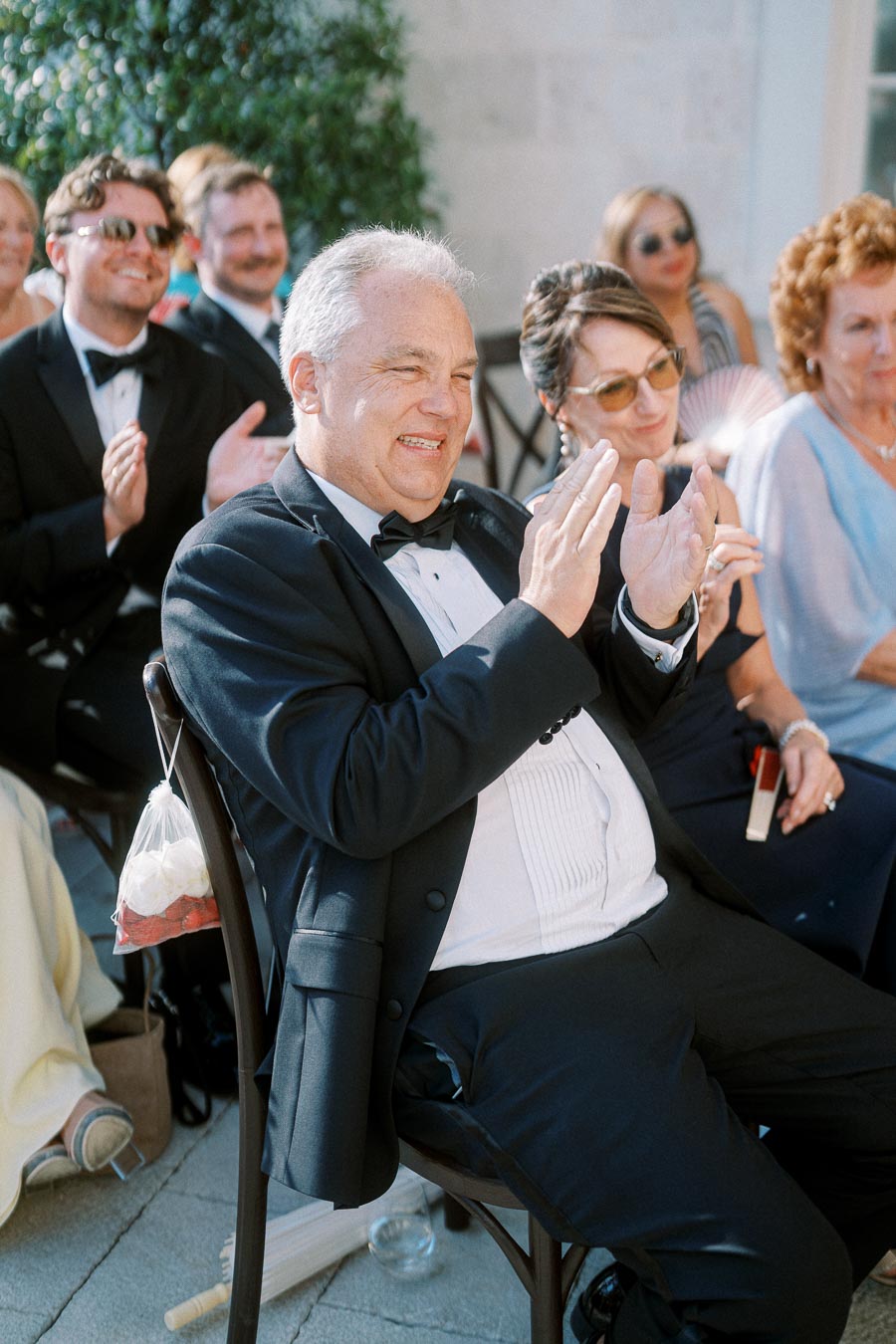 Guests at an outdoor event in formal attire clapping and smiling, with a decorative garden setting in the background.