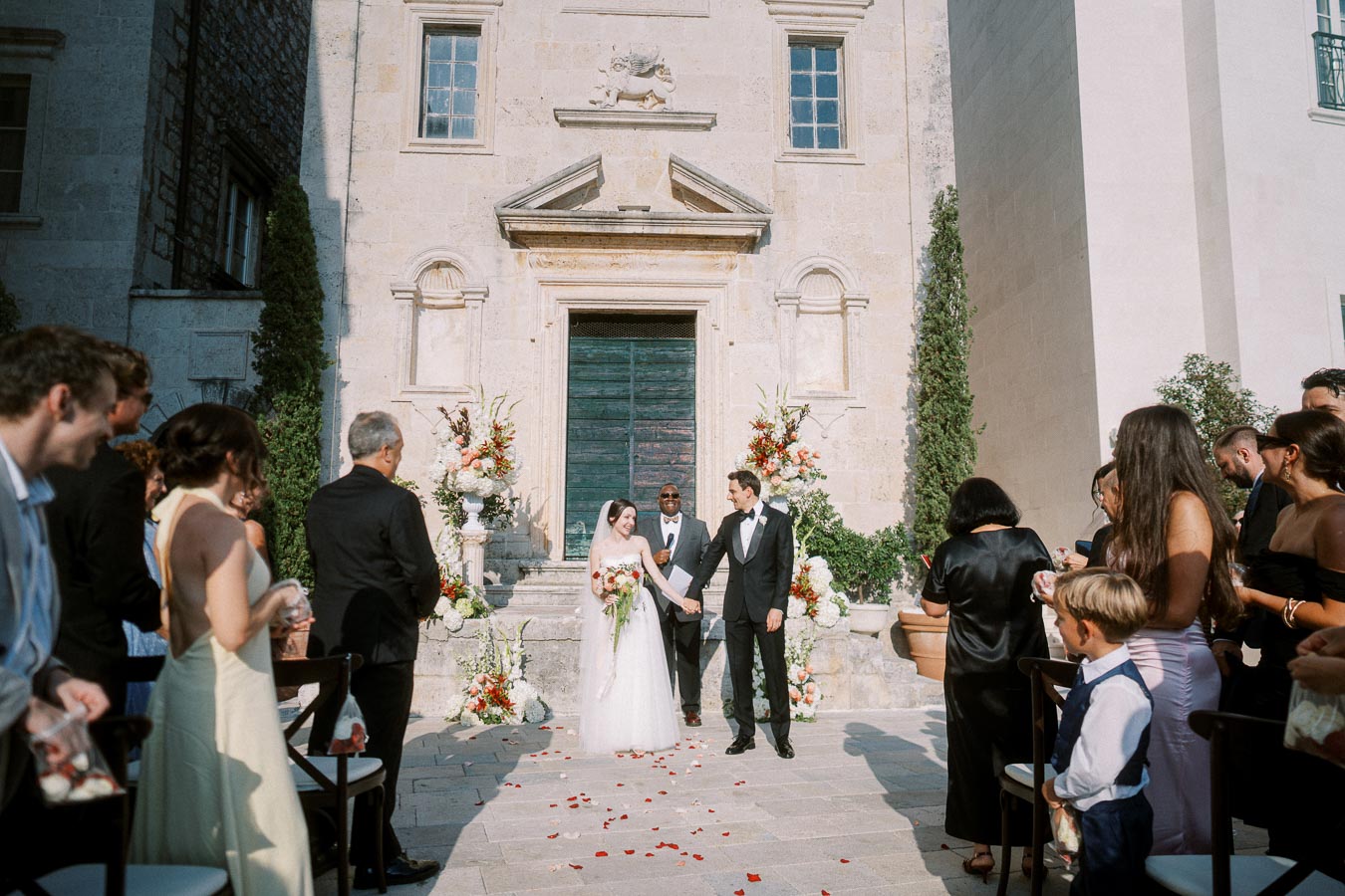 A bride and groom holding hands in front of a historic church during an outdoor wedding ceremony, surrounded by guests and floral decorations.