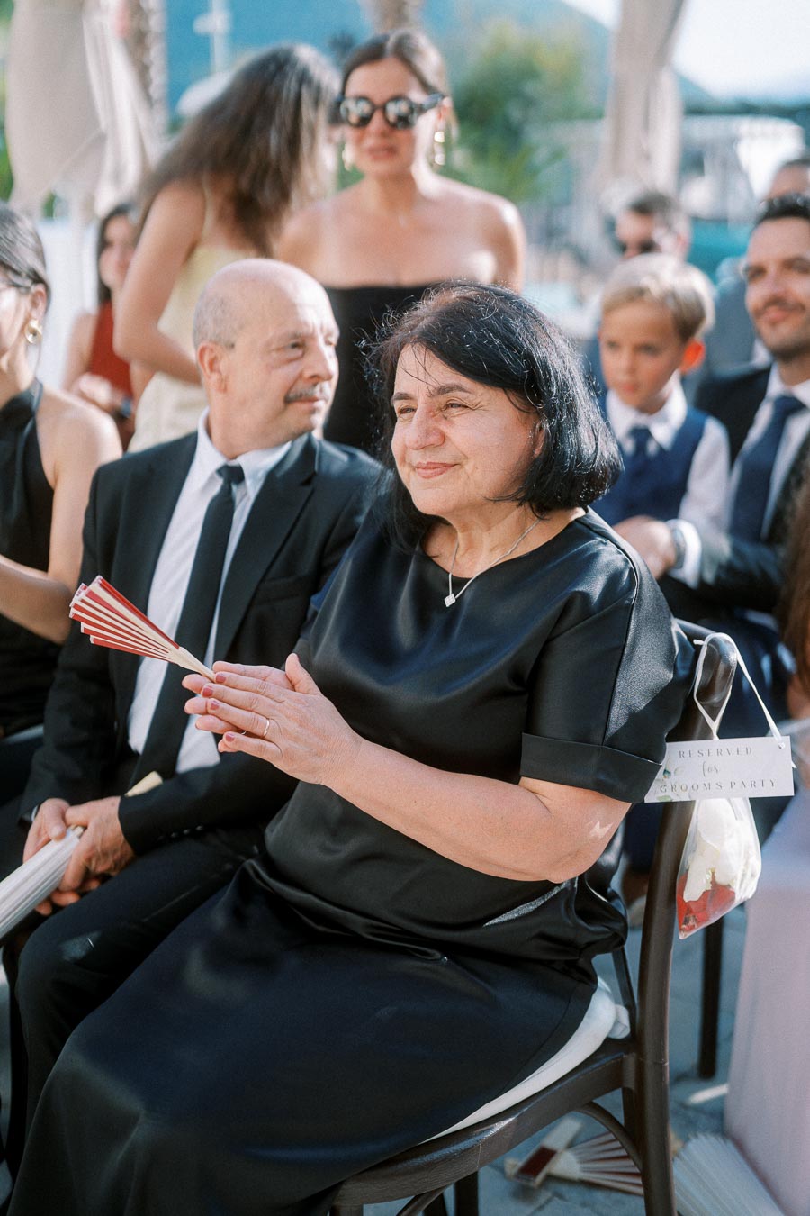 Elderly woman in a black dress clapping at an outdoor formal event, surrounded by well-dressed guests, showcasing a reserved seating sign for the groom's party.