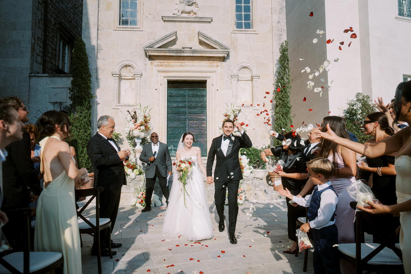 Happy newlywed couple exits a historic church surrounded by cheering guests and flying rose petals in a picturesque wedding ceremony.